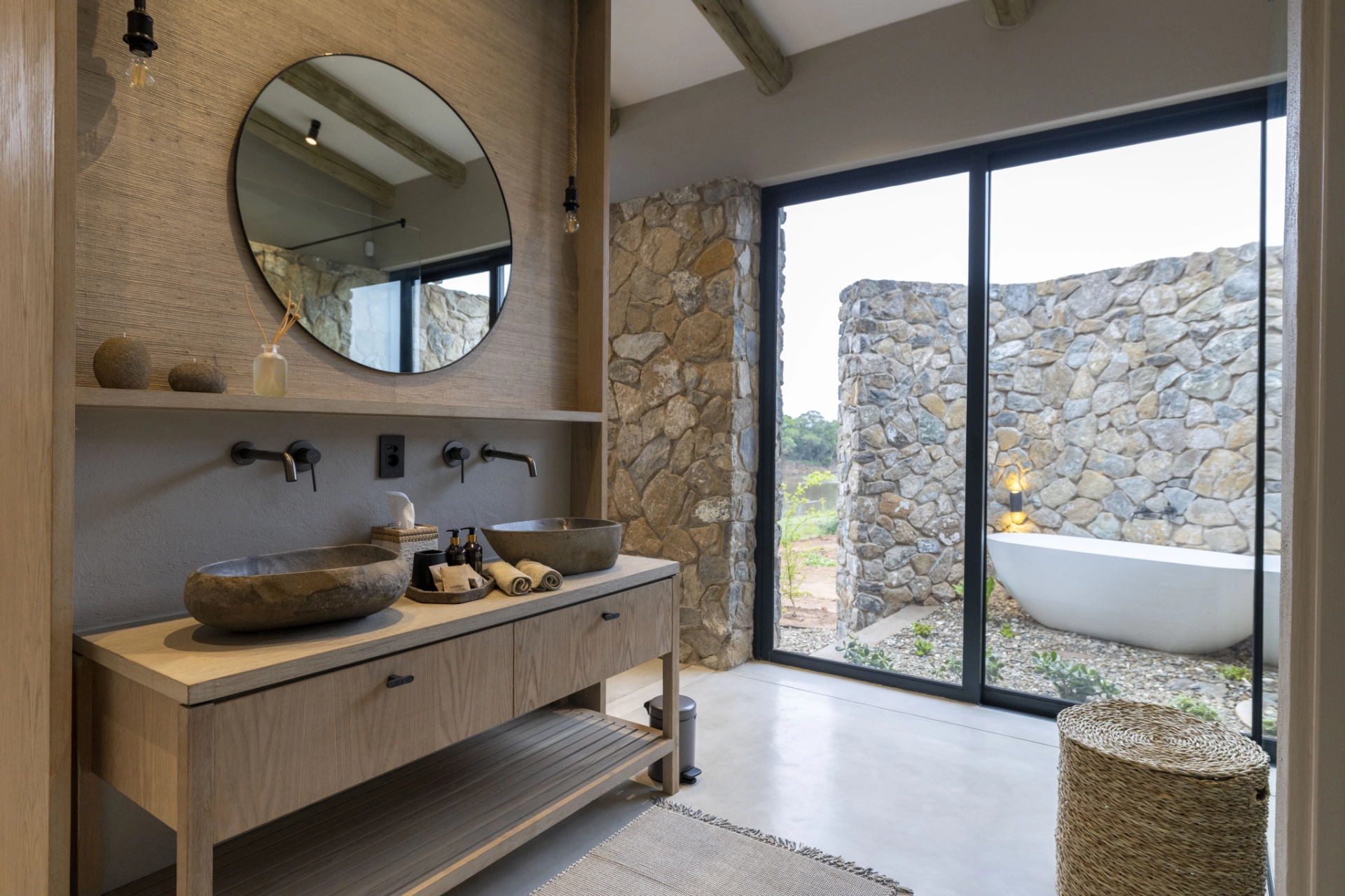 Bathroom with stone basins, round mirror, and large windows opening onto an outdoor bath.