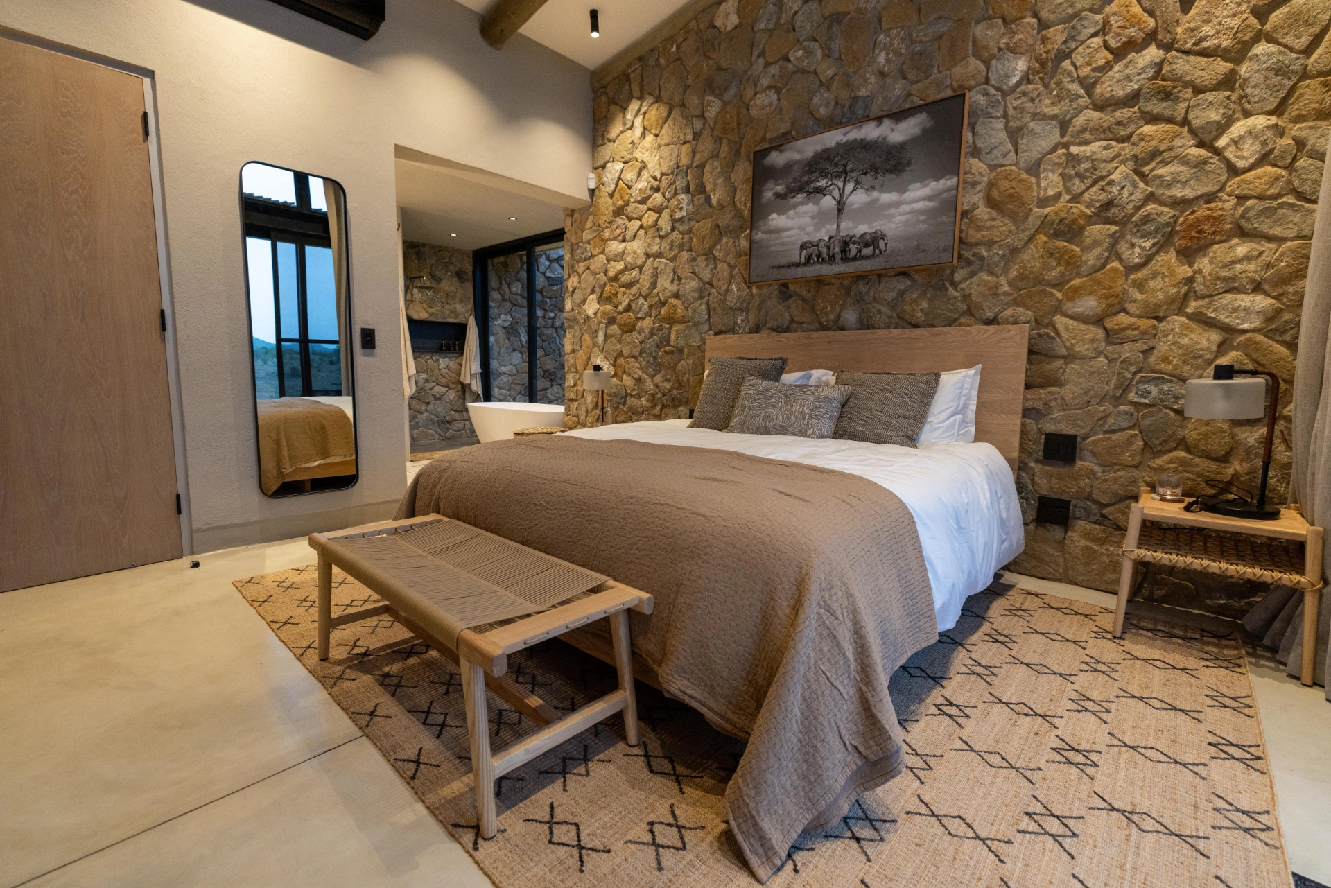 Bedroom with stone accent wall, neutral bedding and wooden furniture inside Izakithi River Villa, overlooking the natural surroundings of Malelane near Kruger Park.