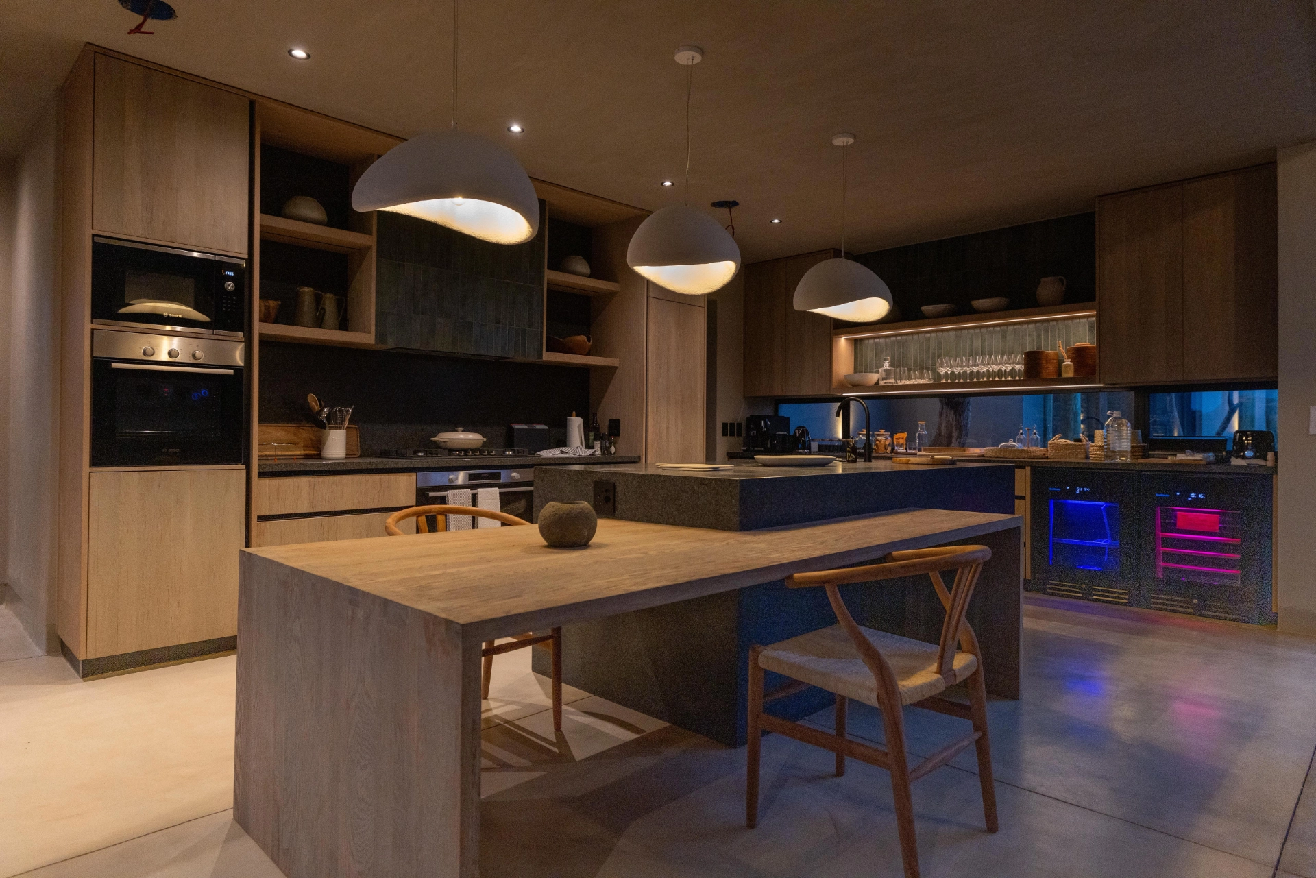 Modern open-plan kitchen at Izakithi River Villa, featuring wooden cabinetry, pendant lights, a large island and a wine fridge, designed with a warm contemporary lodge style